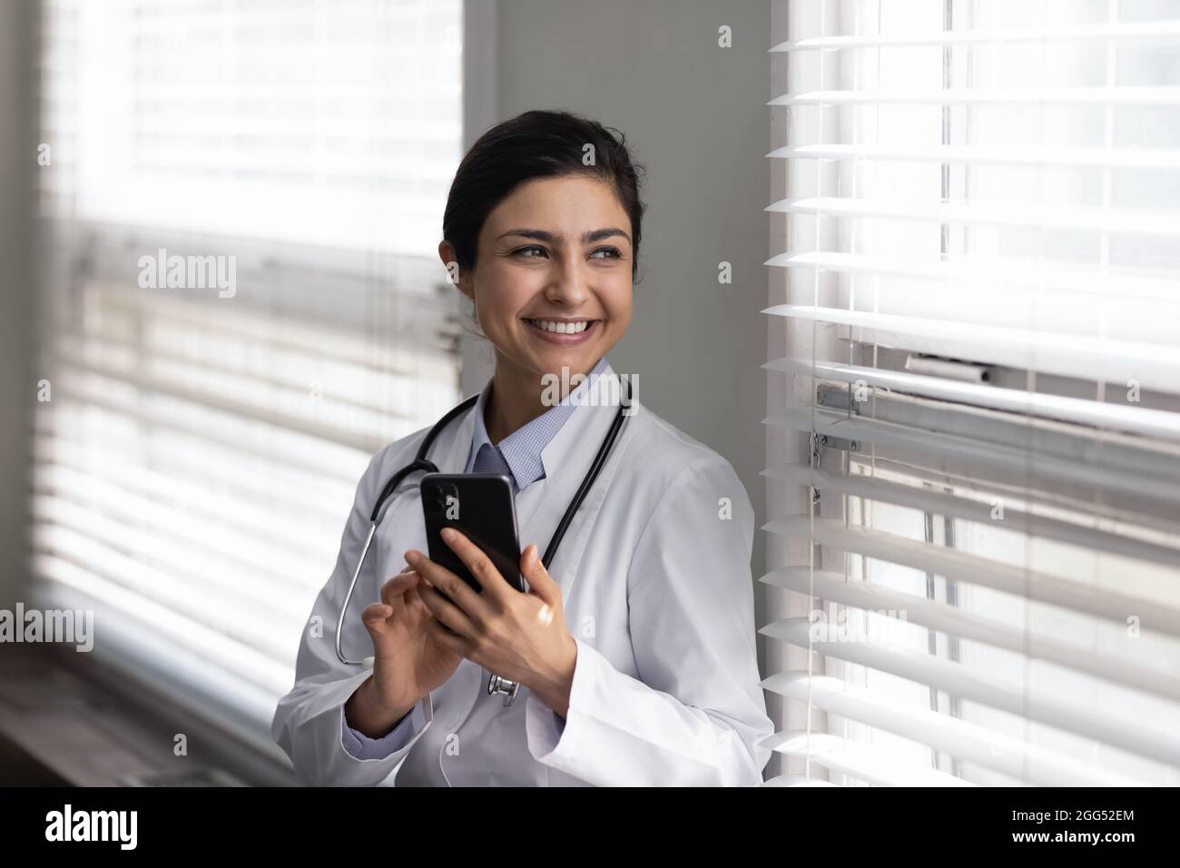 Sorridente medico indiano giovane sognante che tiene il cellulare nelle mani. Foto Stock