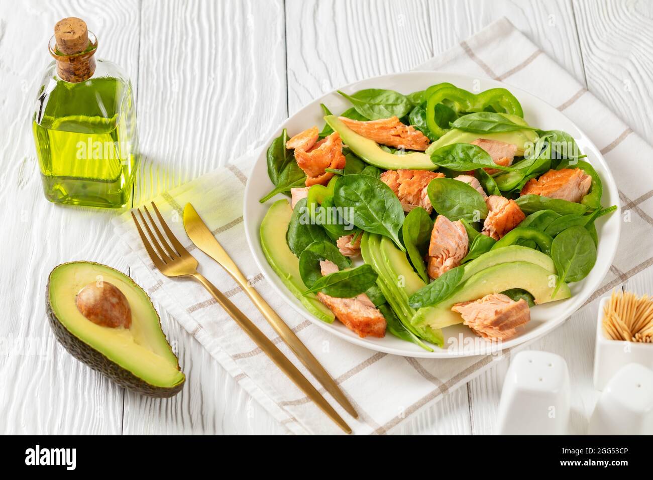 avocado di spinaci al salmone e insalata di pepe verde su un piatto bianco con forchetta dorata e coltello su un tavolo di legno bianco con ingredienti, salute mediterranea Foto Stock