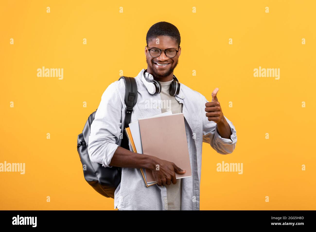 Studente afro-americano che mostra pollice su, posando con zaino, notebook e cuffie sul collo, sfondo giallo Foto Stock