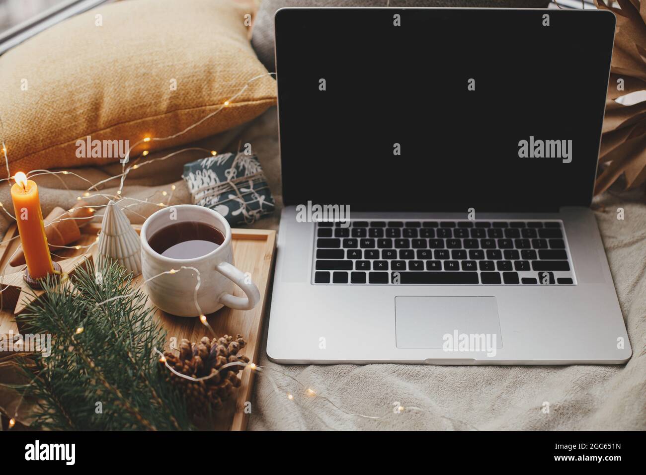 Computer portatile su letto morbido con tazza di tè, luci di natale, stelle, pino, candela e cuscini in camera scandinava. Freelance e blog. Lavoro in remoto e online Foto Stock