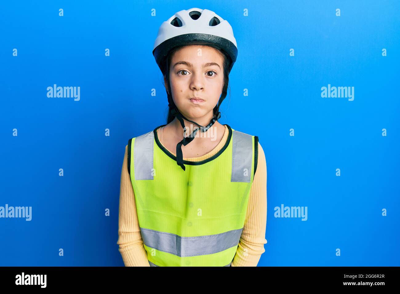 Bella bruna bambina indossando casco da bici e giubbotto riflettente gonfia guance con faccia divertente. Bocca gonfiata con aria, espressione pazza. Foto Stock