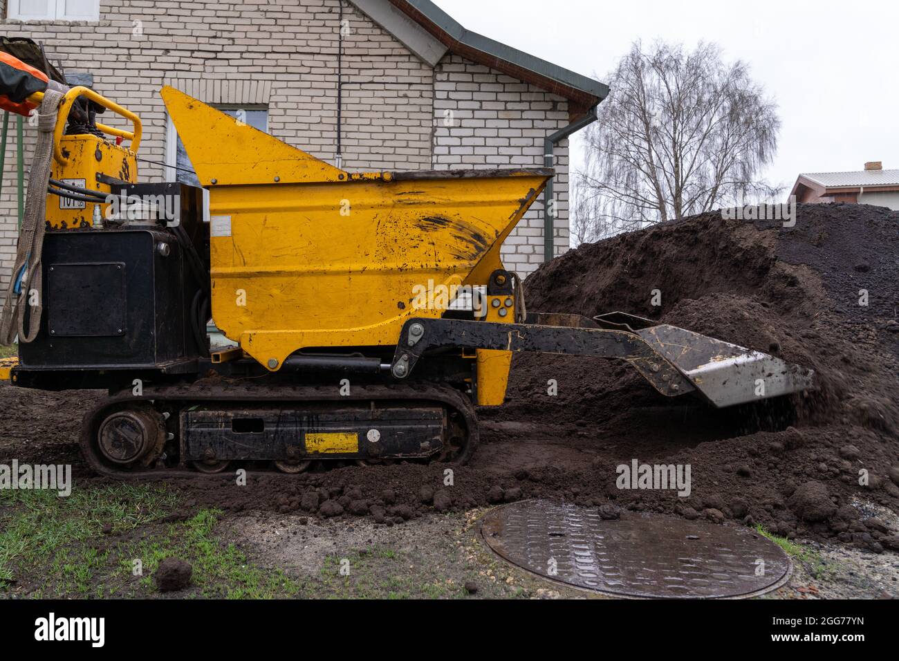 Il piccolo trattore cingolato dumpster muove il terreno fertile, aiutando il giardiniere a impostare il giardino in una giornata di primavera piovosa Foto Stock