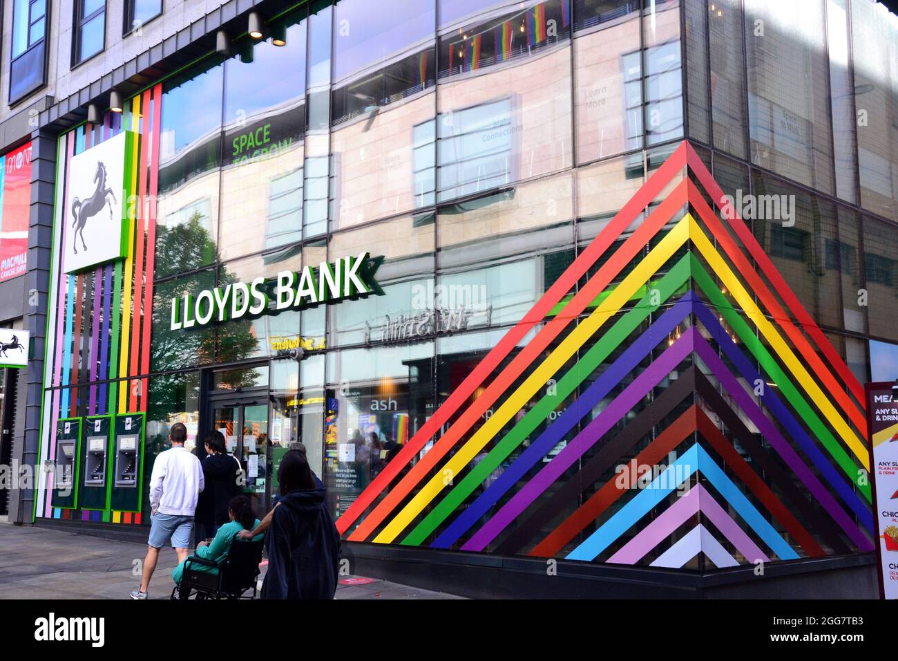 La gente cammina davanti a una filiale della banca Lloyds in Market Street, Manchester, Inghilterra, regno unito, che è decorato in colori arcobaleno per il week-end LGBTQ Pride di Manchester. Foto Stock