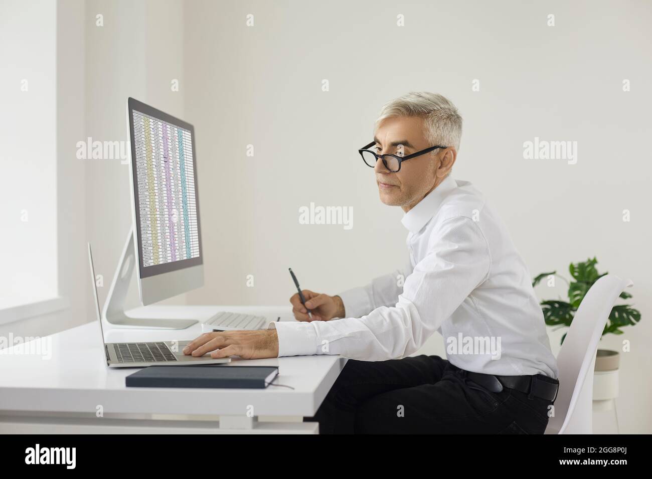 Commercialista o uomo d'affari seduto alla scrivania dell'ufficio e che lavora su computer portatili e desktop Foto Stock