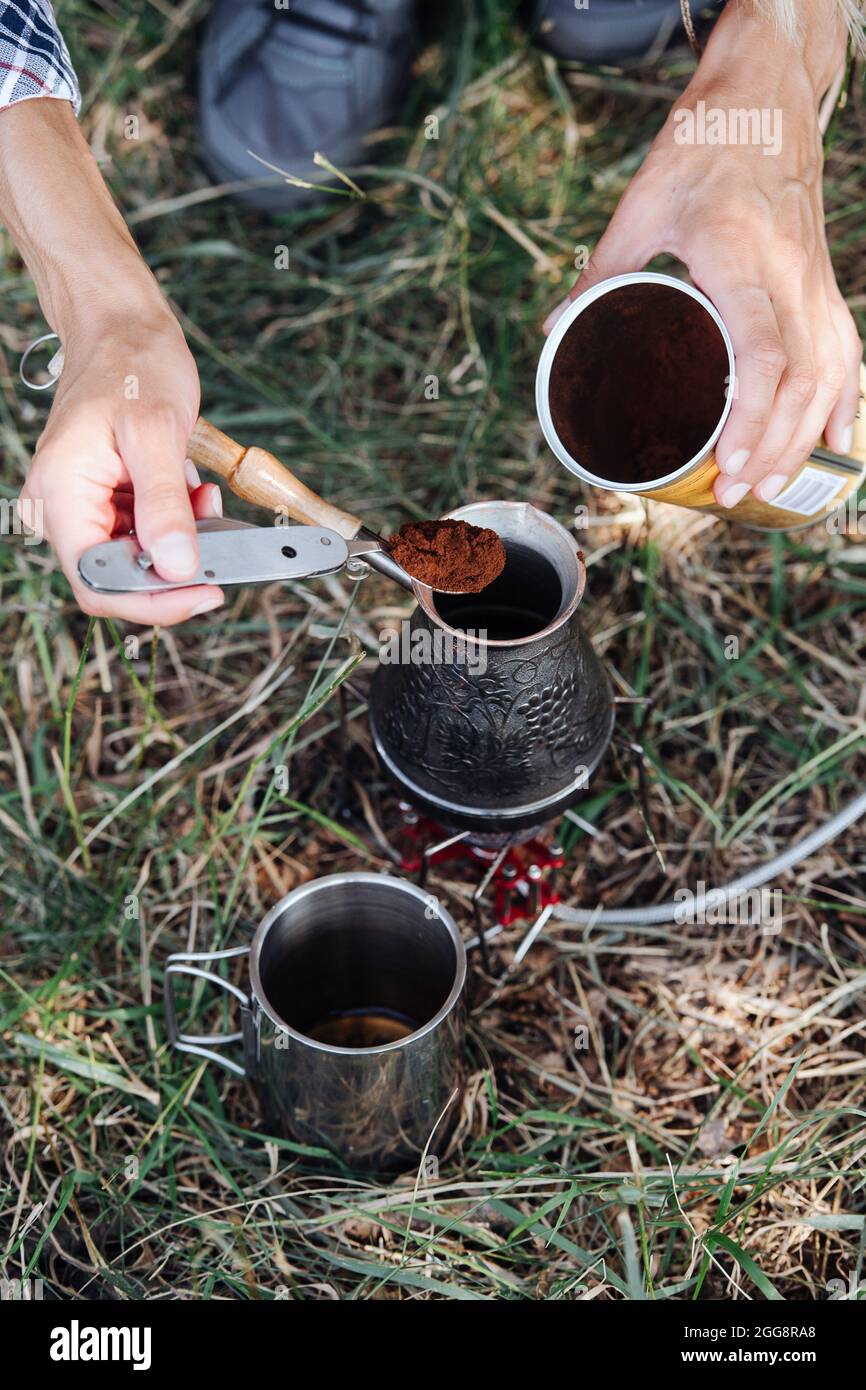 Le mani aggiungono la polvere di caffè in una cezve con un cucchiaio pieghevole di metallo. Su un prato durante il campeggio. Foto Stock
