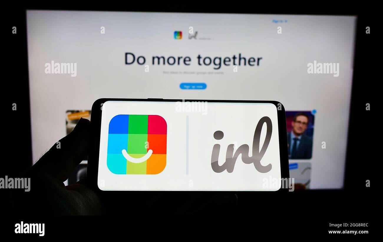 Persona che tiene uno smartphone con il logo del social network statunitense IRL (Get Together Inc.) sullo schermo di fronte al sito Web. Mettere a fuoco sul display del telefono. Foto Stock