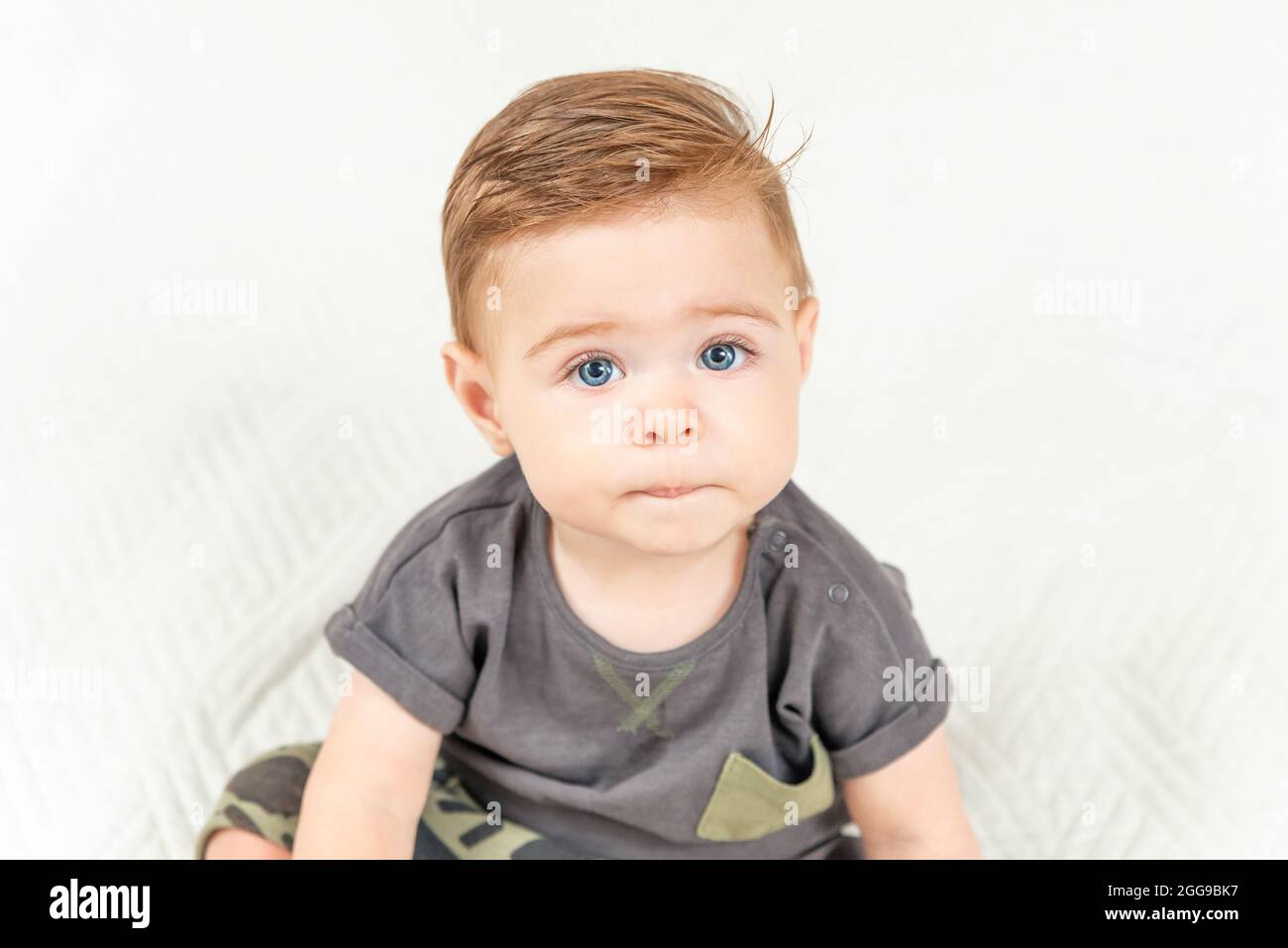 Ritratto del bambino carino seduto sul letto in camera, sfondo bianco, spazio copia. bambino di 1 anno. Foto Stock