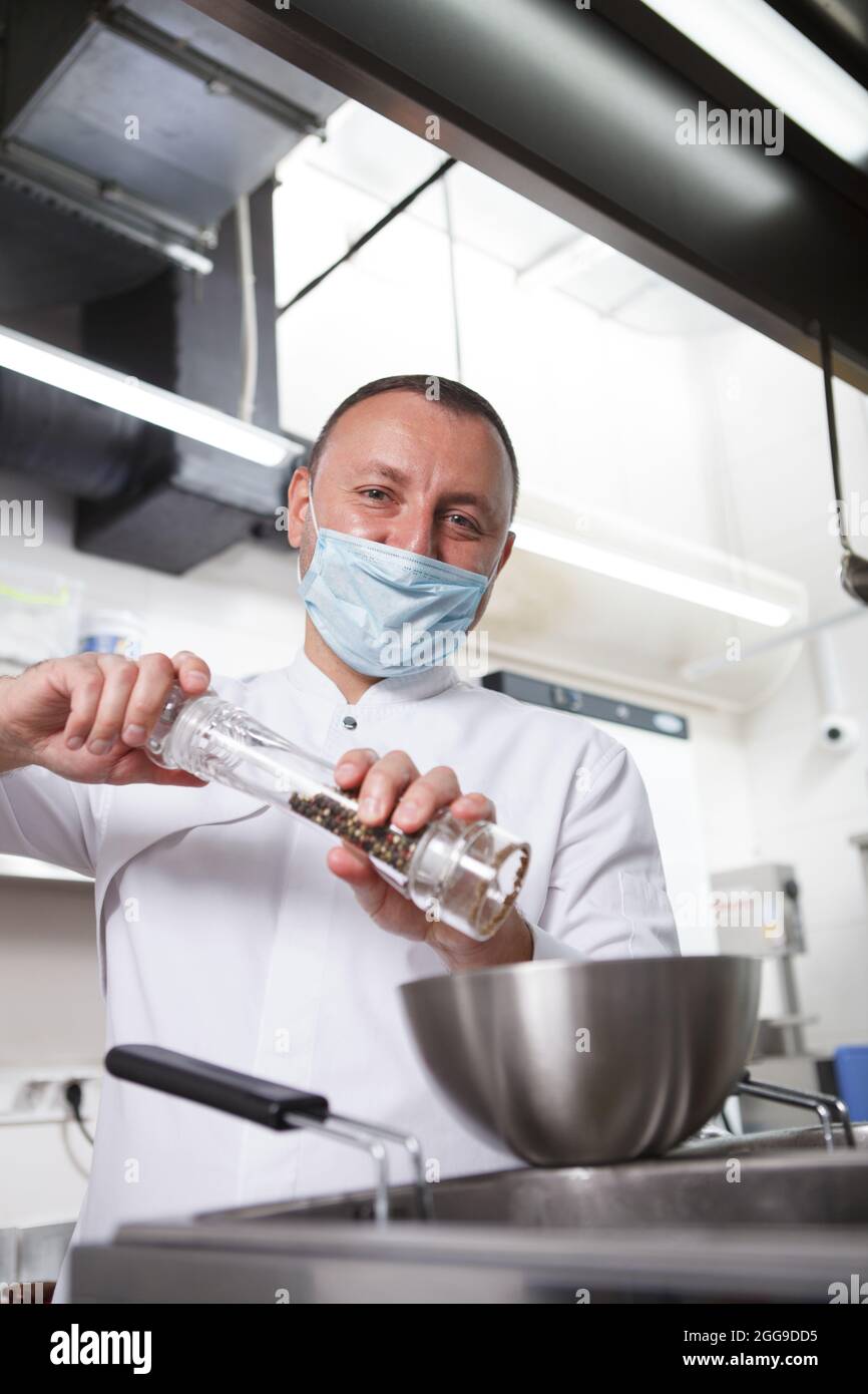 Ritratto verticale di uno chef in maschera medica salatura pasto preparato al ristorante cucina Foto Stock
