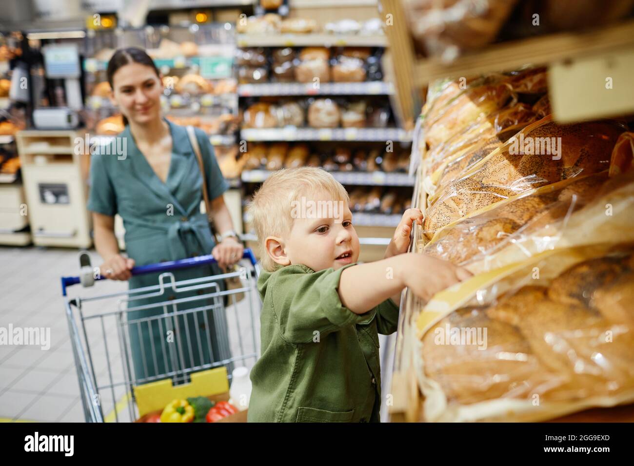 Figlio carino in camicia casual prendendo il pane dalla mensola mentre facendo gli acquisti con la madre in negozio Foto Stock