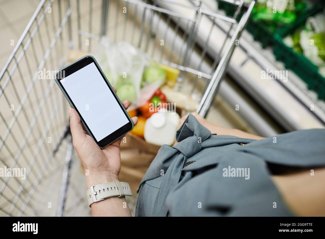 Sopra la vista di una donna incinta irriconoscibile in piedi al carrello e controllare le note sul telefono mentre comprano i prodotti nel punto vendita Foto Stock
