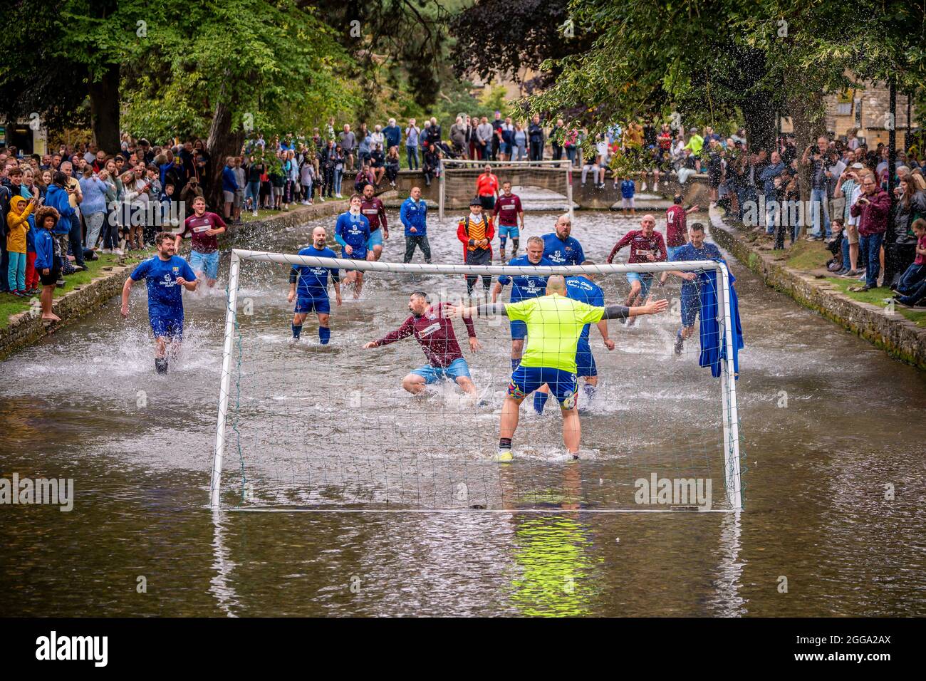 Le folle fiaccano il fiume Windrush mentre i calciatori di Bourton Rovers creano un tuffo mentre combattono per la palla durante la tradizionale partita di football fluviale annuale nel villaggio di Cotswolds, Bourton-in-the-Water, Gloucestershire, dove la scoreline è caduta a un 1-1 pareggio. Data foto: Lunedì 30 agosto 2021. Foto Stock