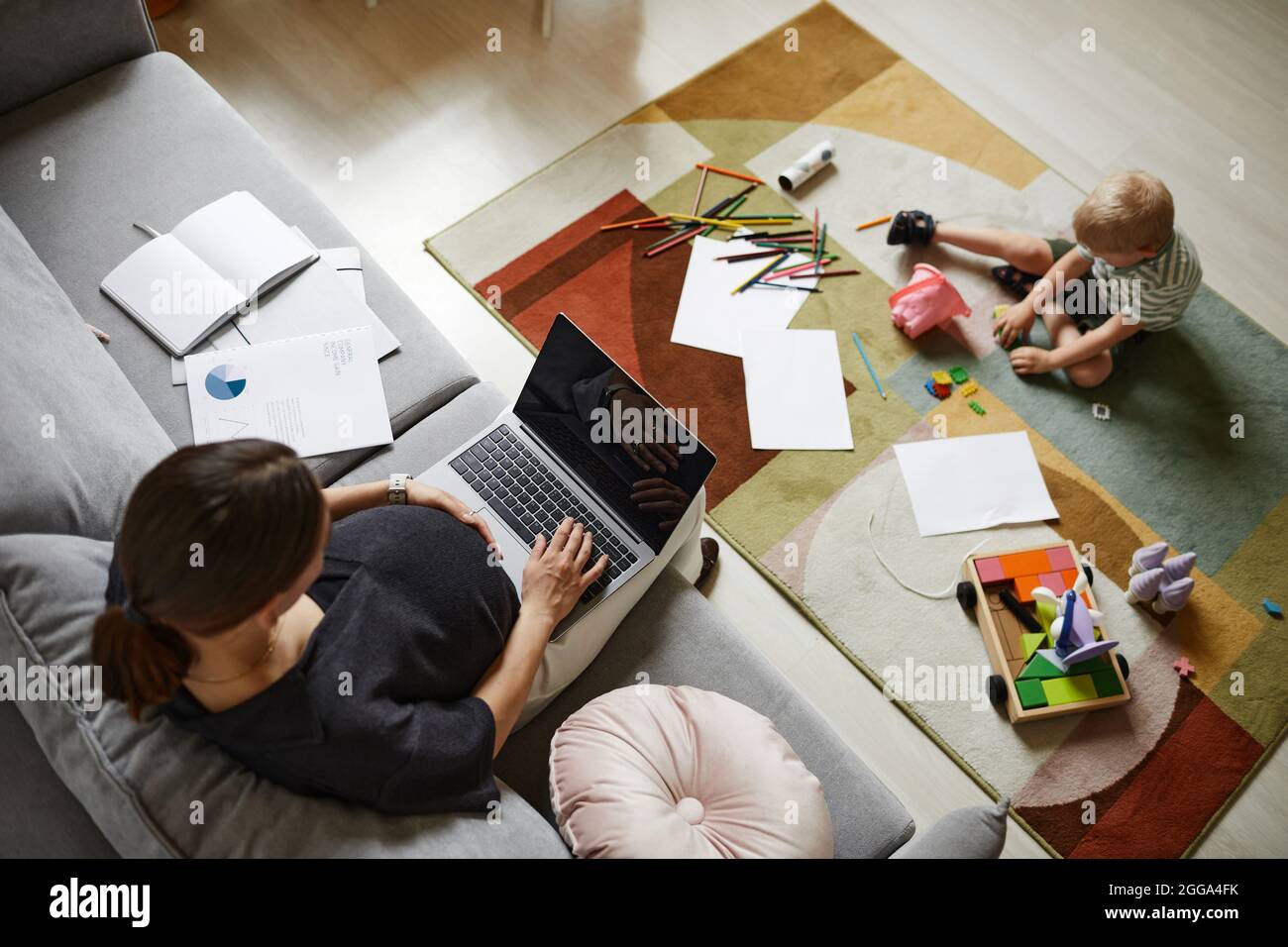 Sopra il viiew della madre incinta occupata usando il laptop mentre suo figlio gioca con i giocattoli sul tappeto nel soggiorno Foto Stock