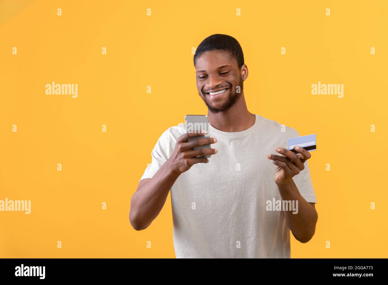 Concetto di portafoglio online. Felice ragazzo nero che tiene smartphone e carta di credito, in piedi su sfondo giallo, spazio libero Foto Stock
