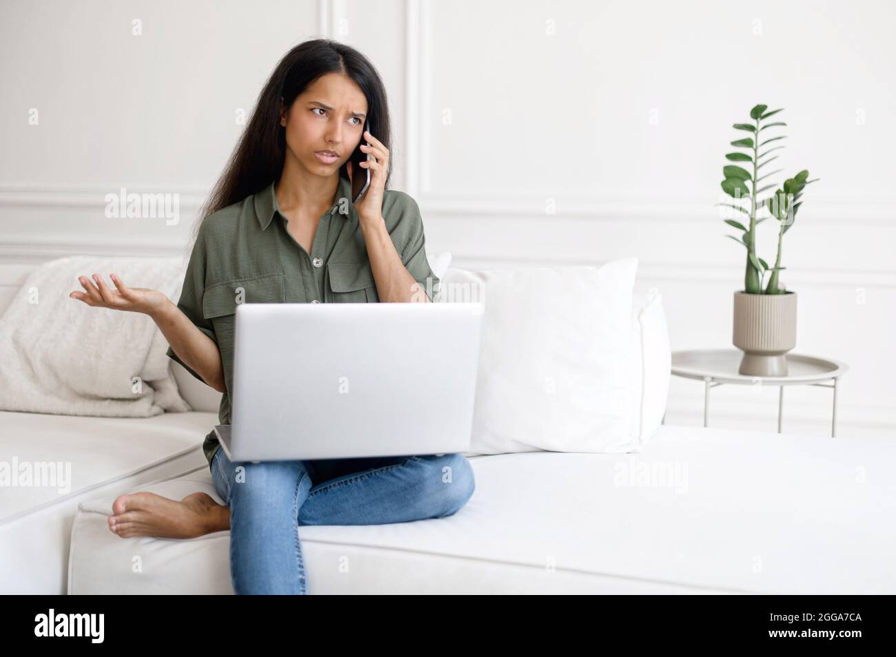 Donna indiana irritata che parla al telefono mentre si siede sul divano a casa e si utilizza un computer portatile. Turbato e pensivo freelance femmina parlare sullo smartphone, non capire qualcosa correttamente Foto Stock