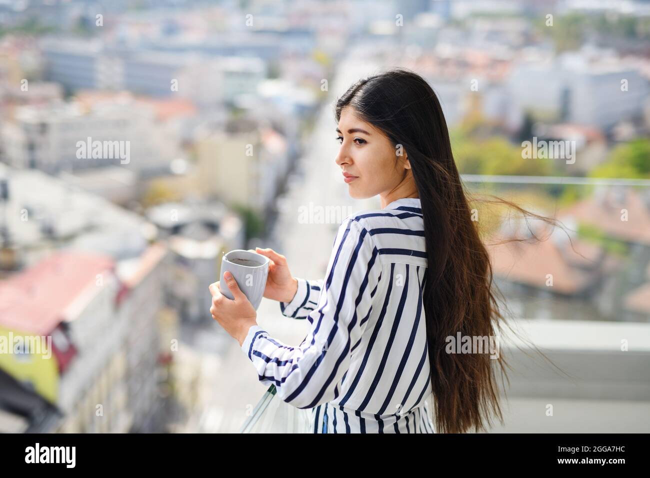 Ritratto di giovane donna con caffè in piedi sul balcone all'aperto in città, guardando il paesaggio urbano. Foto Stock