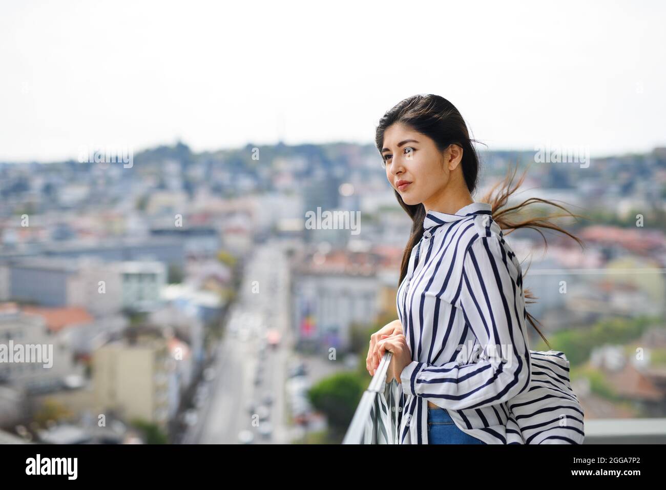 Ritratto di giovane donna in piedi sul balcone all'aperto in città, guardando il paesaggio urbano. Foto Stock
