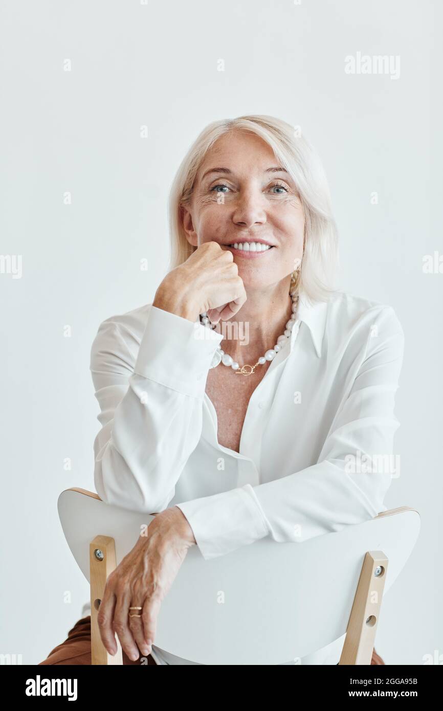 Ritratto verticale di donna matura elegante sorridente seduto su sedia dietro su sfondo bianco Foto Stock
