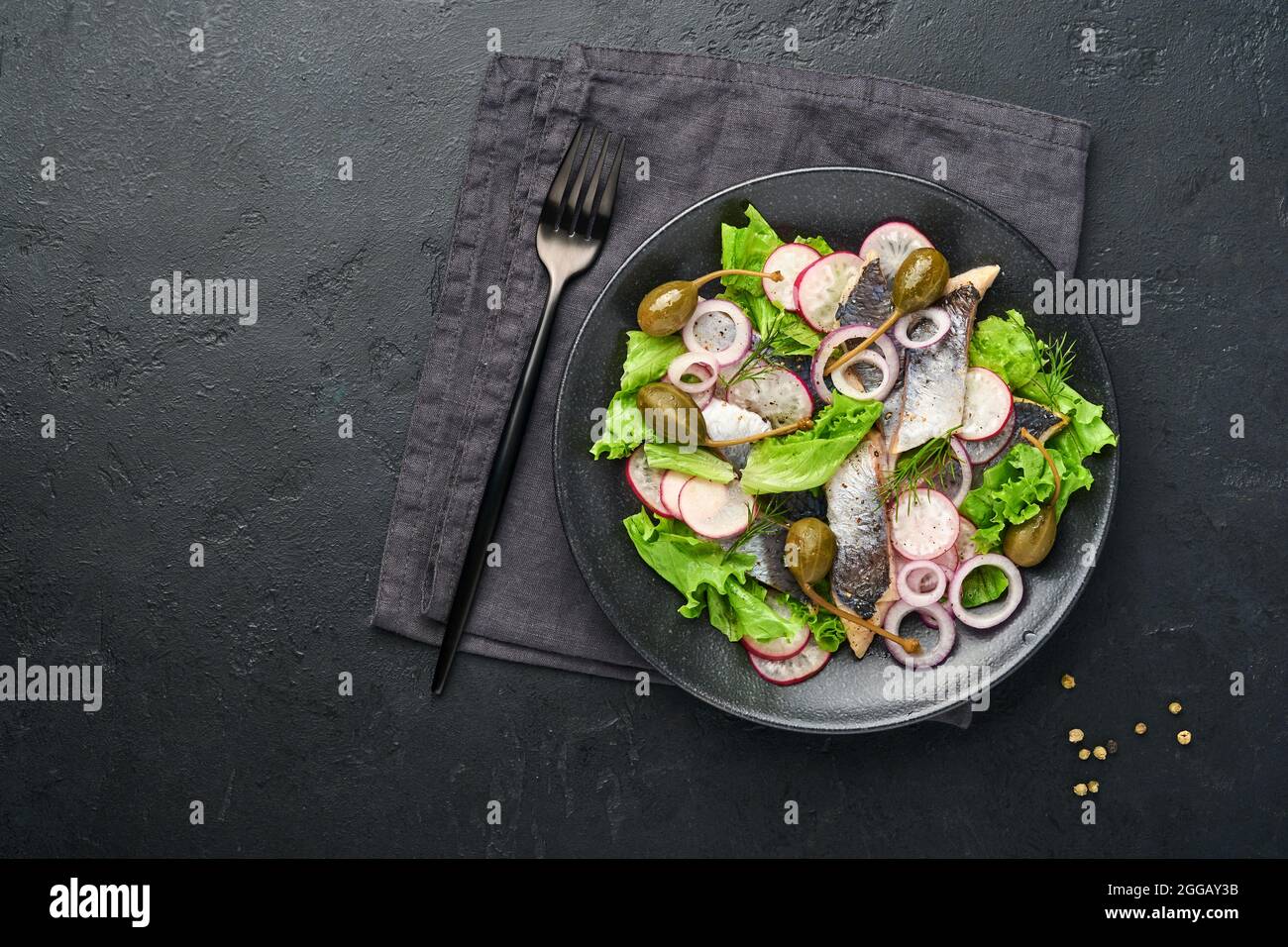 Aringhe salate con spezie, capperi, erbe e insalata di cipolla su piatto nero su sfondo scuro con spazio copia. Pesce marinato a fette. Cibo con u sano Foto Stock