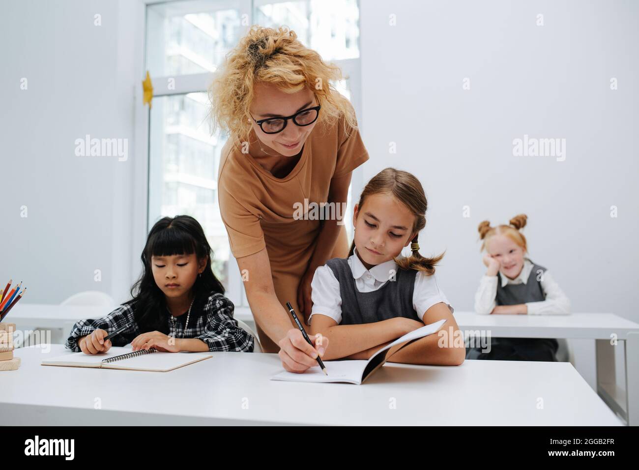 Insegnante che aiuta una studentessa seduta dietro una scrivania, scrivendo nel suo notebook. Vista frontale. Foto Stock