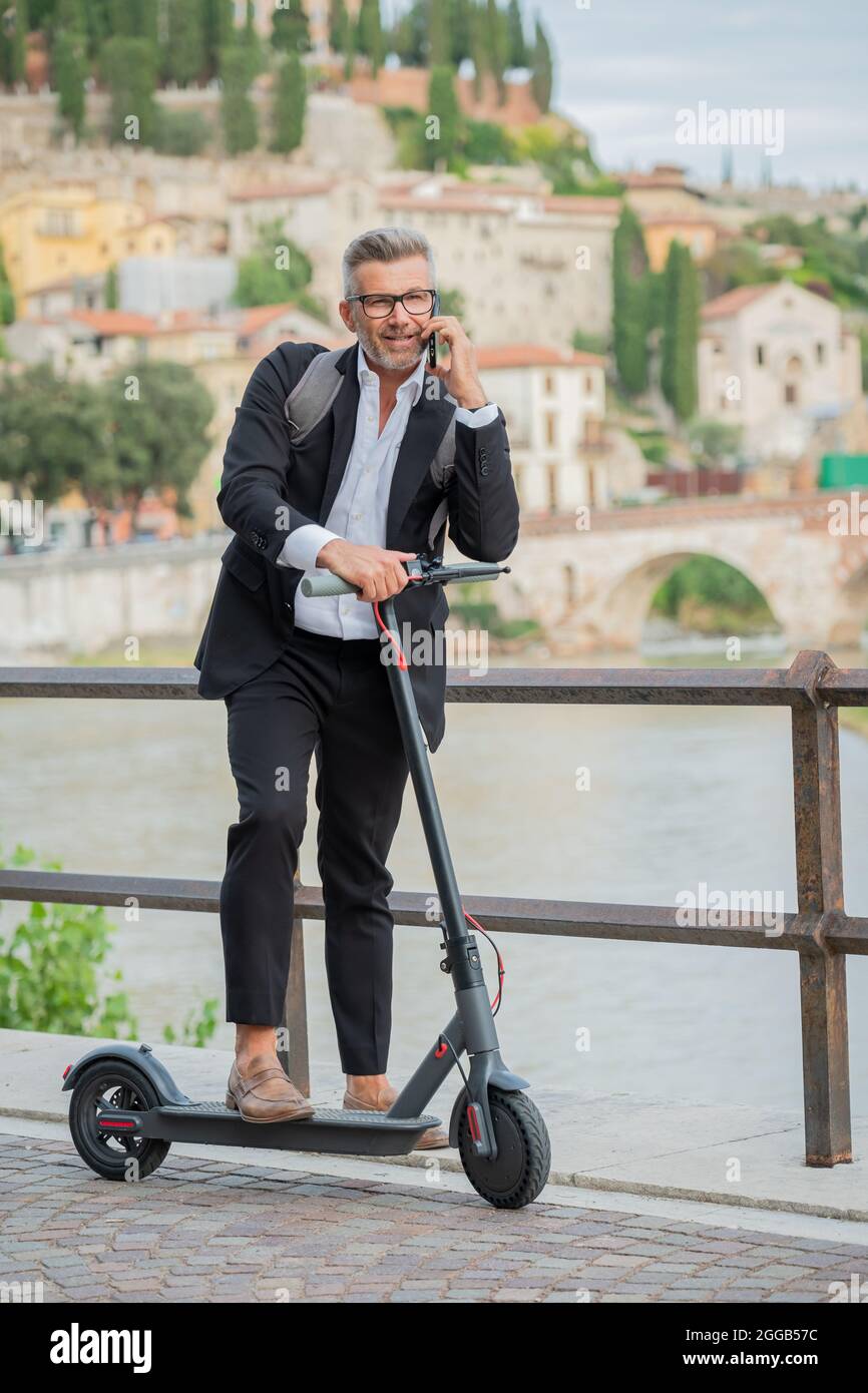 Uomo d'affari con abito elegante in piedi sullo scooter elettrico mentre parla al telefono. Imprenditore che guida lo scooter e risponde al telefono Foto Stock