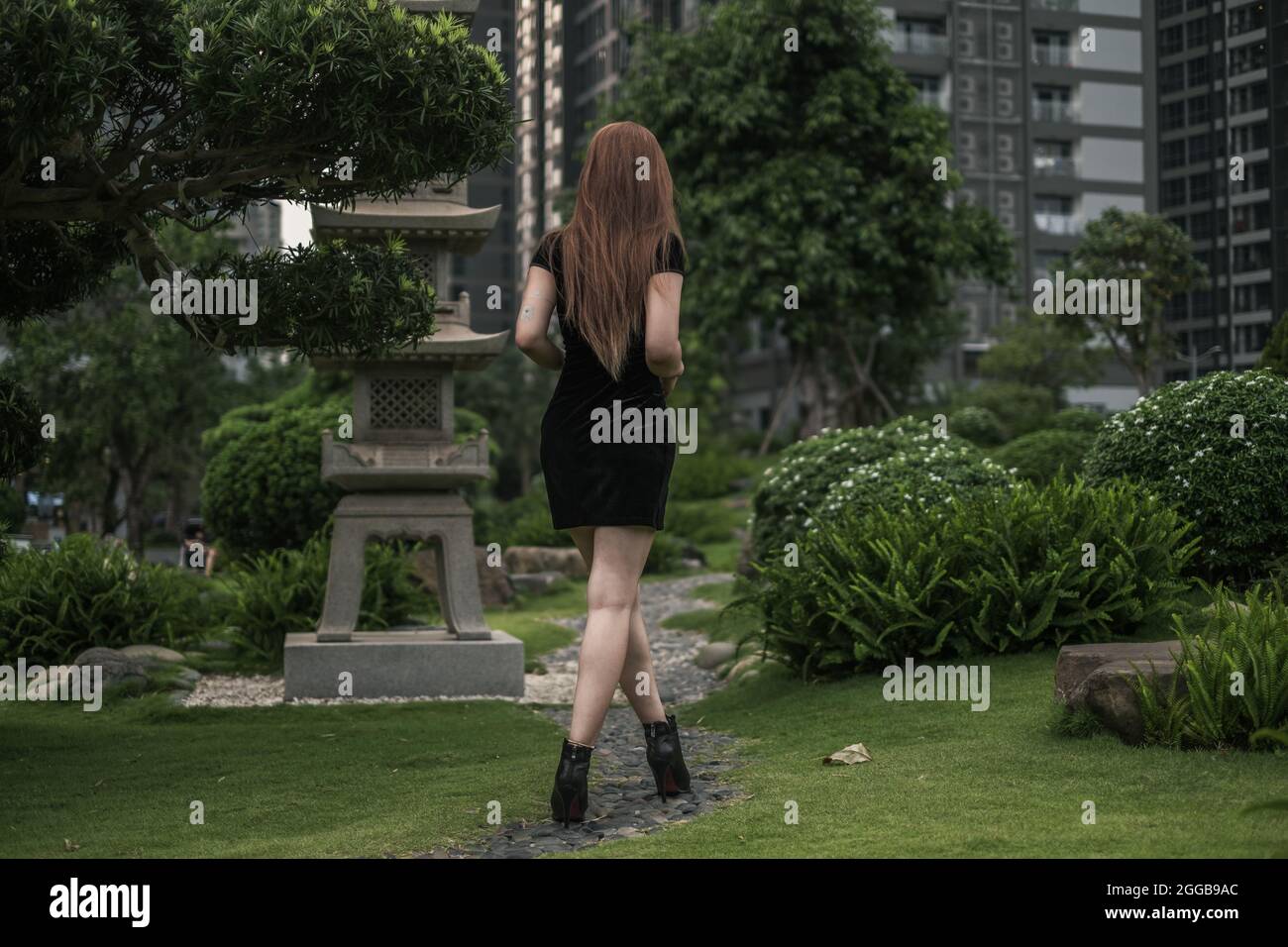 Bella giovane donna asiatica in mini abito nero a piedi nel parco. Capelli lunghi. Bella postura. Vista laterale posteriore. Immagine dal retro. Foto Stock