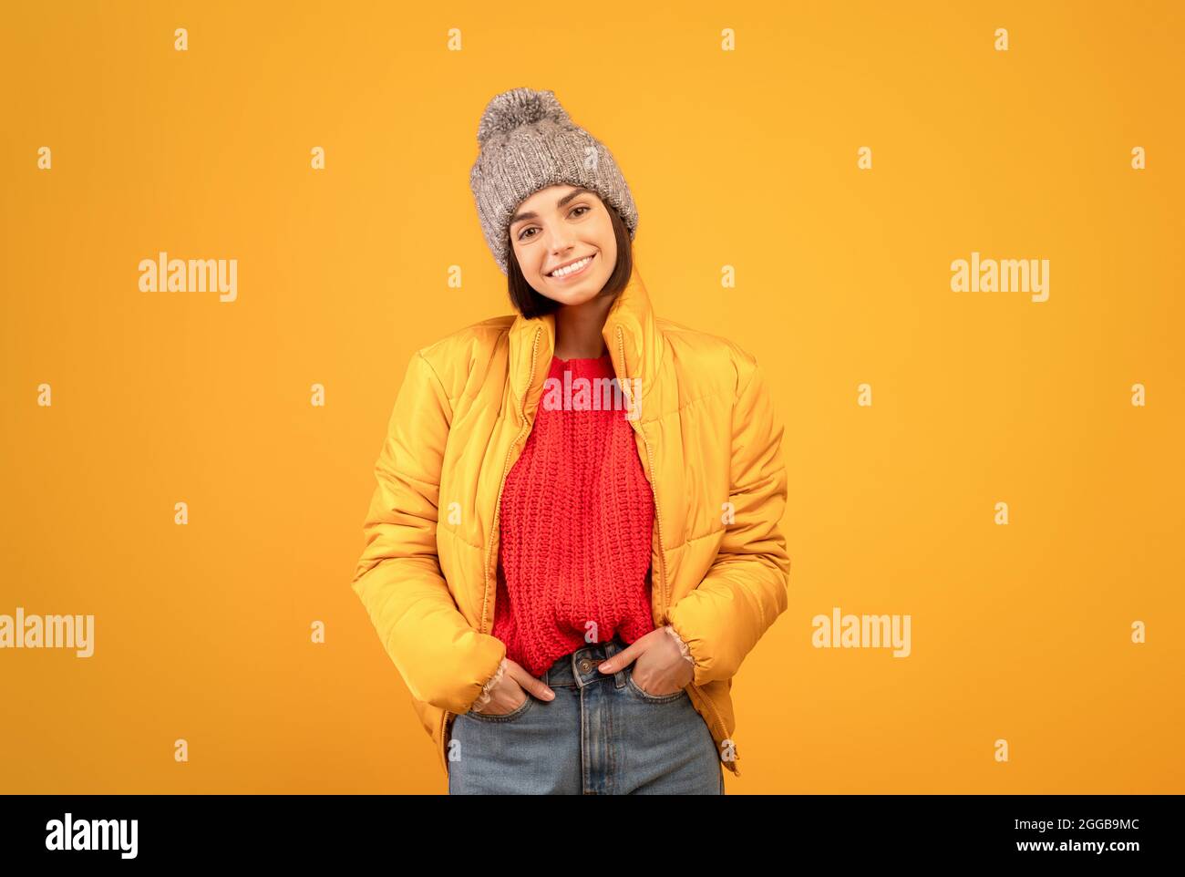Donna felice in giacca calda, maglione a maglia e cappello che tiene le mani in tasca e sorridendo alla macchina fotografica, sfondo arancione Foto Stock