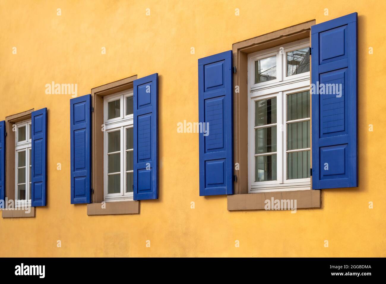 tre finestre bianche con davanzali in pietra arenaria in fila con persiane blu scuro e una parete della casa dipinta di giallo arancio Foto Stock