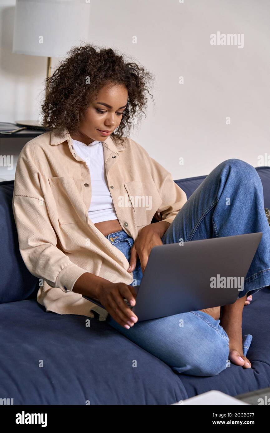 Studentessa afroamericana seduta su un morbido divano a casa digitando su un computer portatile. Foto Stock