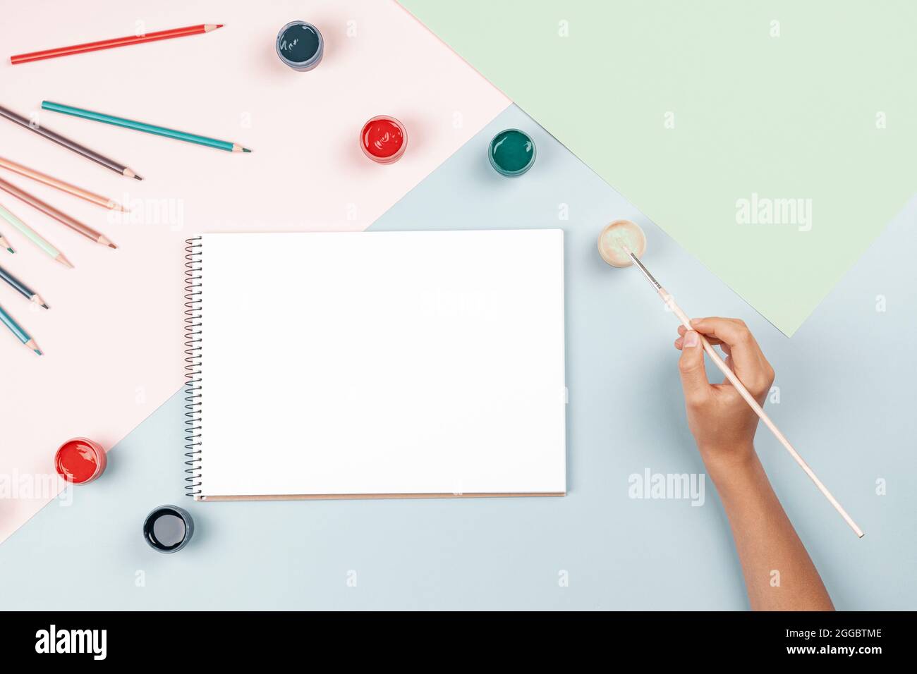 Vista dall'alto sulla mano del bambino con pittura a pennello in quaderno di bozze d'arte. Forniture di opere d'arte creative su sfondo pastello. Torna a scuola Foto Stock