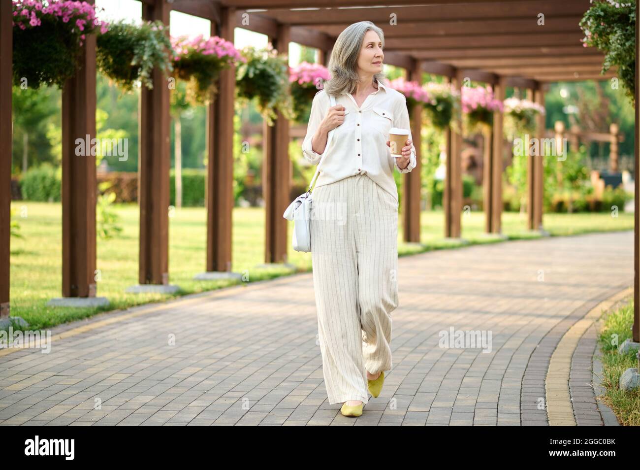 Donna dai capelli grigi con caffè in un parco fiorito Foto Stock