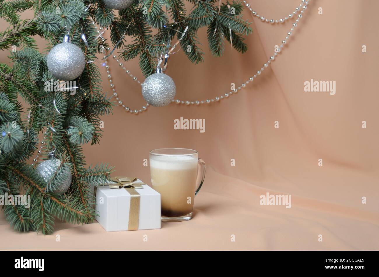 Una composizione di Capodanno fatta di rami di un albero di Natale, decorata con palline d'argento, una scatola regalo, una tazza di vetro con caffè e latte sul dorso Foto Stock