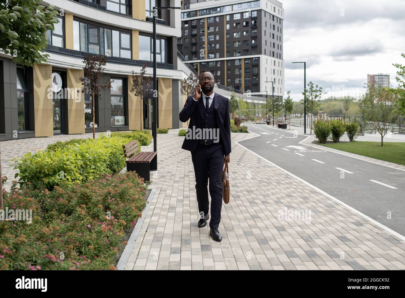 Uomo d'affari sicuro nero in abbigliamento formale che parla sul telefono cellulare mentre si sposta lungo un edificio moderno Foto Stock
