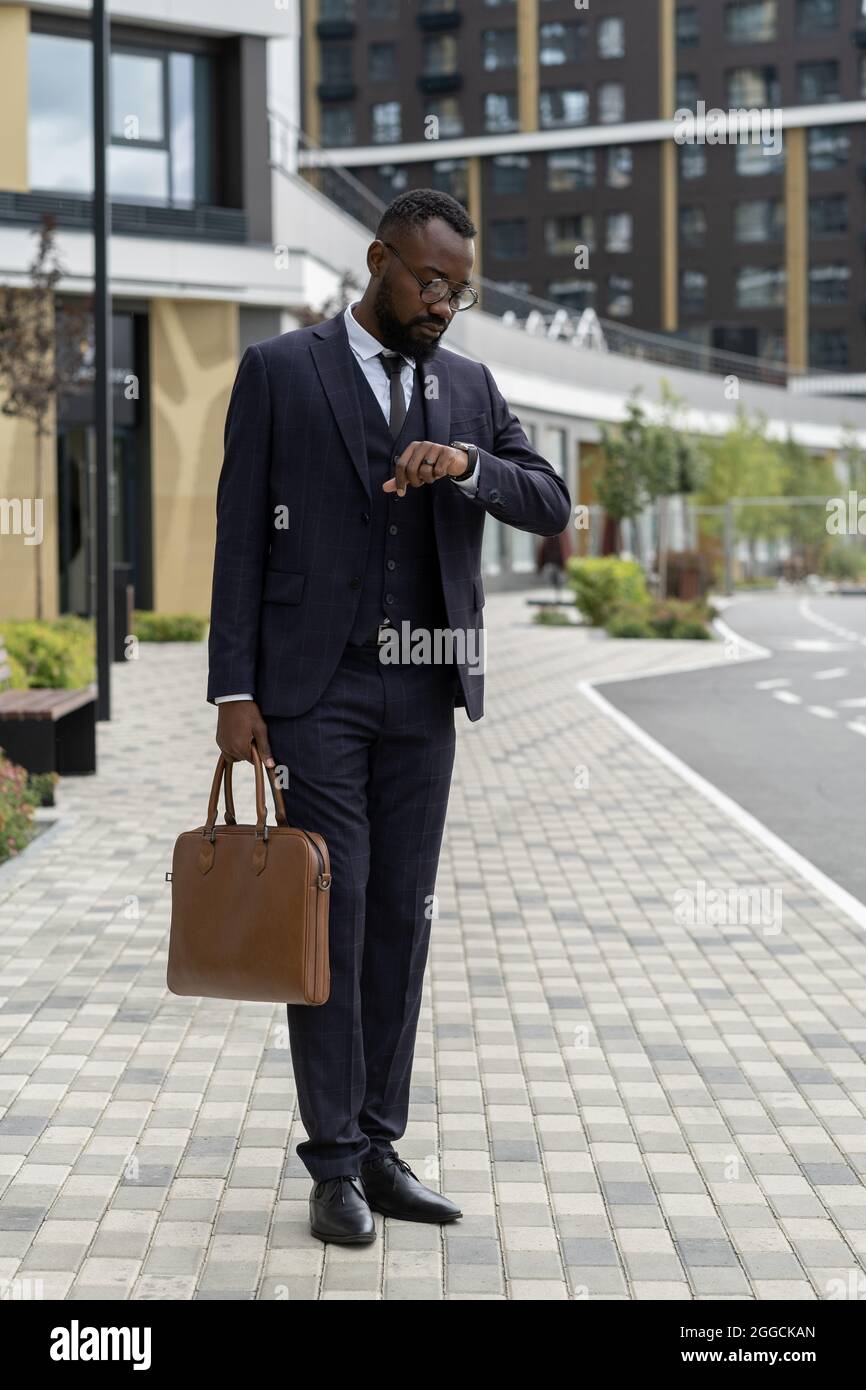 Uomo d'affari elegante con borsa che guarda il orologio da polso mentre si trova in posizione urbana e in attesa di un collega Foto Stock