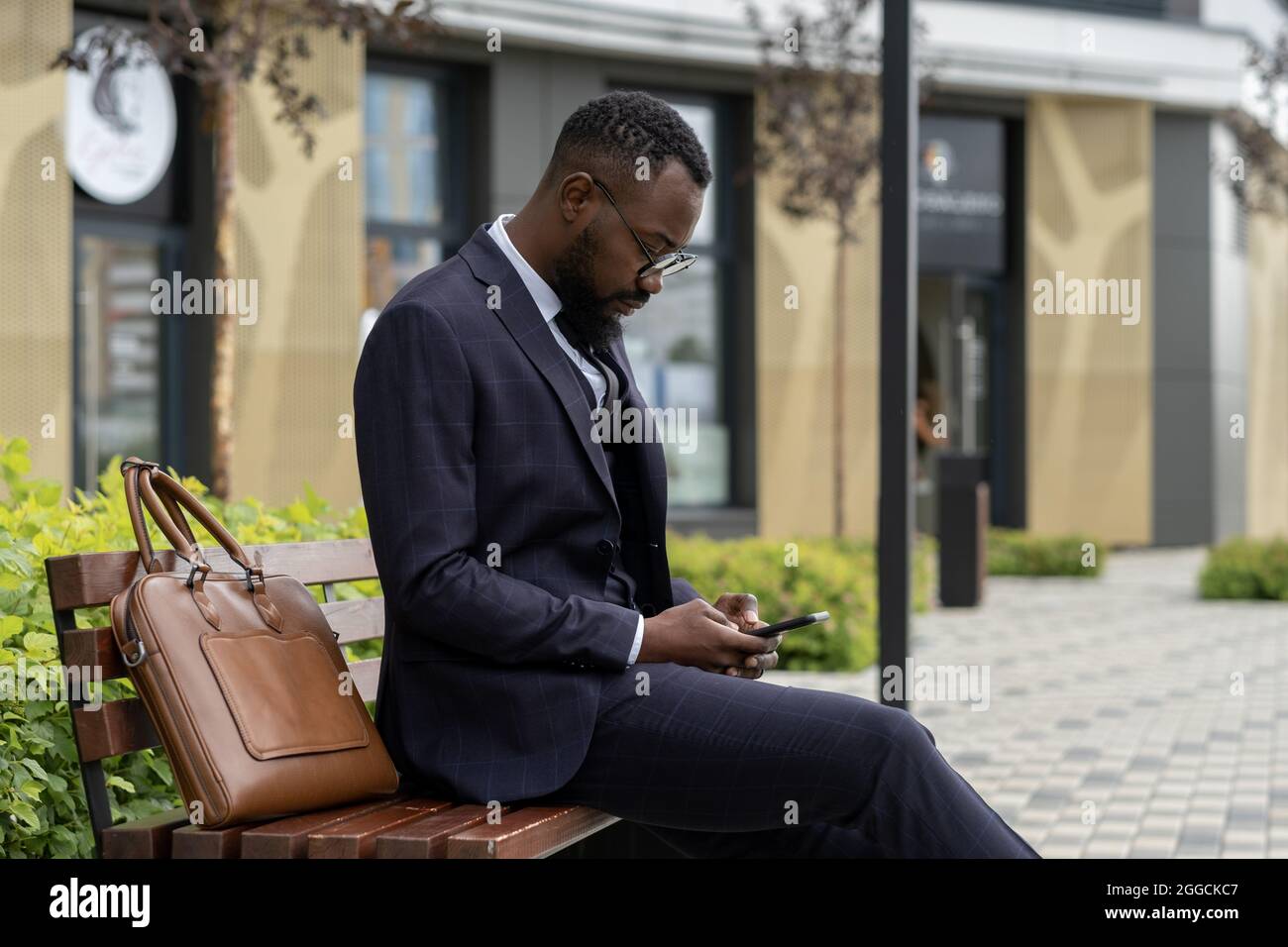 Giovane uomo d'affari di etnia africana che texting in smartphone mentre si riposa sul banco in ambiente urbano Foto Stock