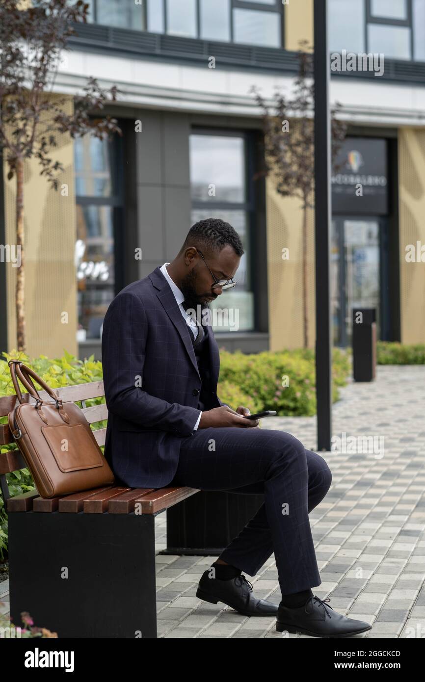 Un uomo d'affari africano serio in tuta che scorre nello smartphone mentre si siede sulla panchina Foto Stock