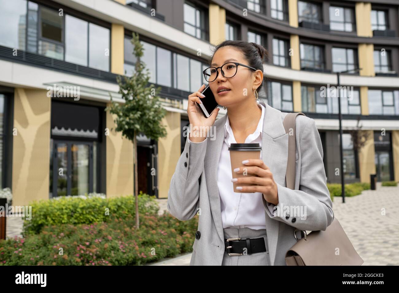 Giovane donna d'affari asiatica con smartphone per orecchio e bere in mano in movimento in ambiente urbano Foto Stock