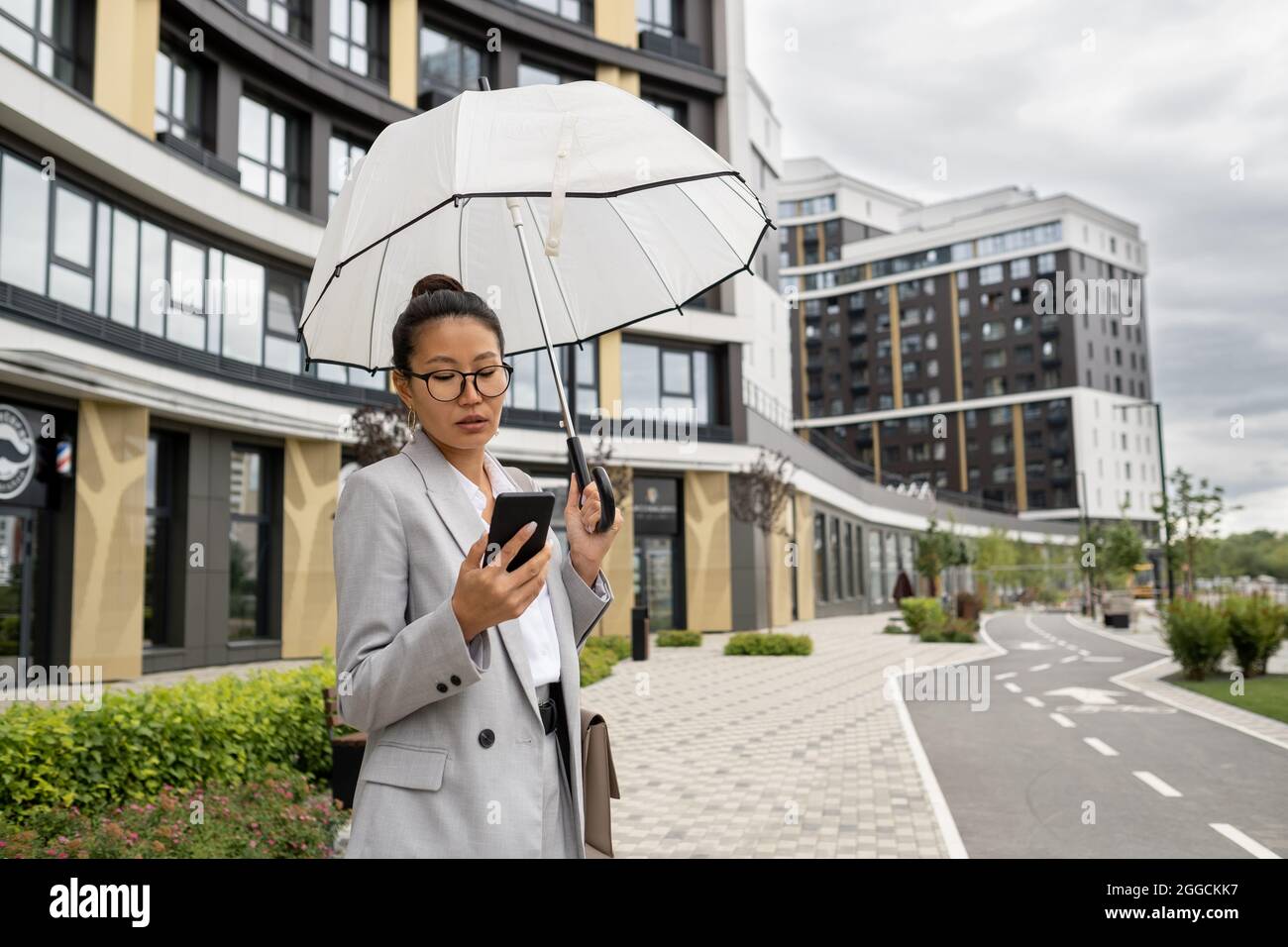 Donna d'affari asiatica con ombrello scorrevole in smartphone in ambiente urbano in giorno di pioggia Foto Stock