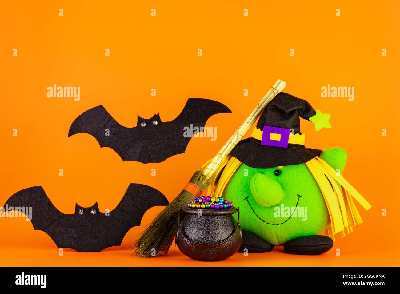 Bambola di strega verde, con il suo cauldron e il broomstick insieme a due graziosi pipistrelli su sfondo giallo arancione. Concetto di tema di Halloween. Foto Stock