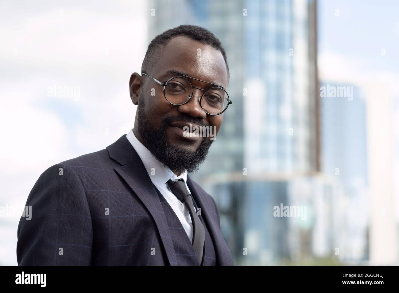 Felice giovane uomo d'affari africano in abbigliamento formale e occhiali guardando la macchina fotografica in ambiente urbano Foto Stock