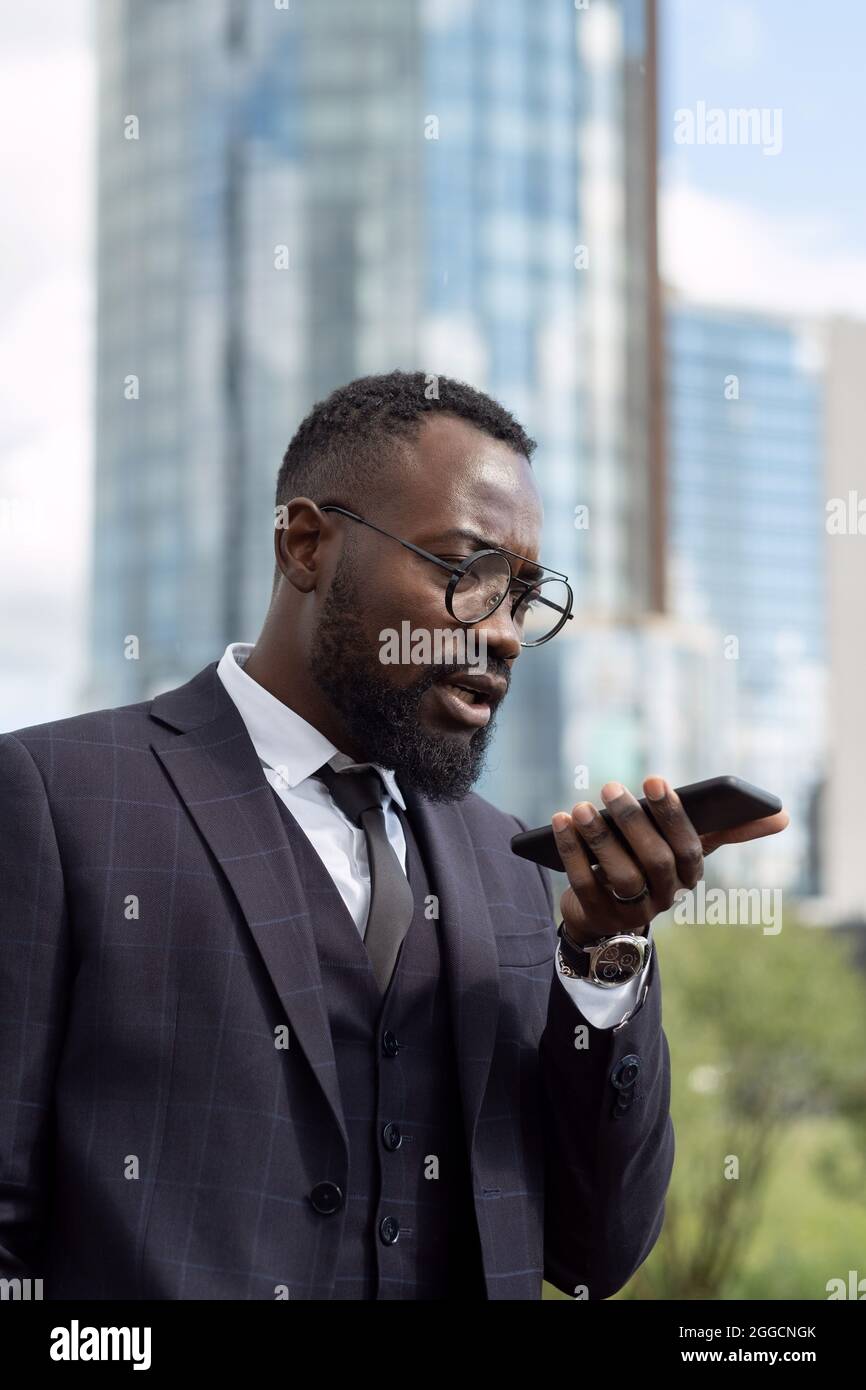 Uomo d'affari contemporaneo che registra i messaggi vocali mentre tiene lo smartphone dalla parte inferiore del viso Foto Stock