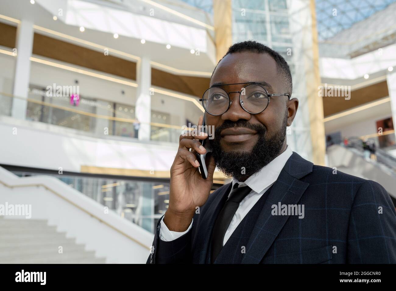 Felice giovane uomo d'affari africano che tiene smartphone per orecchio mentre parla con il cliente all'interno di un grande centro affari Foto Stock