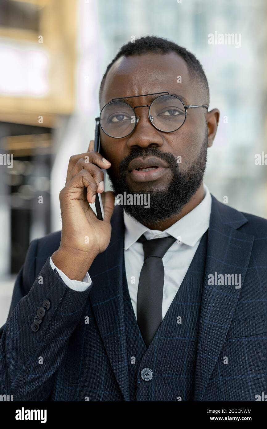 Giovane uomo d'affari africano contemporaneo in vestito e occhiali tenendo smartphone per le orecchie durante le telefonate Foto Stock