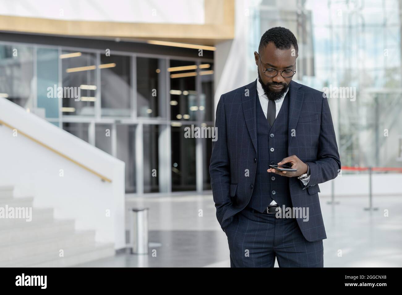 Giovane mediatore afroamericano in elegante abito testando in smartphone all'interno di un grande centro d'ufficio contemporaneo Foto Stock