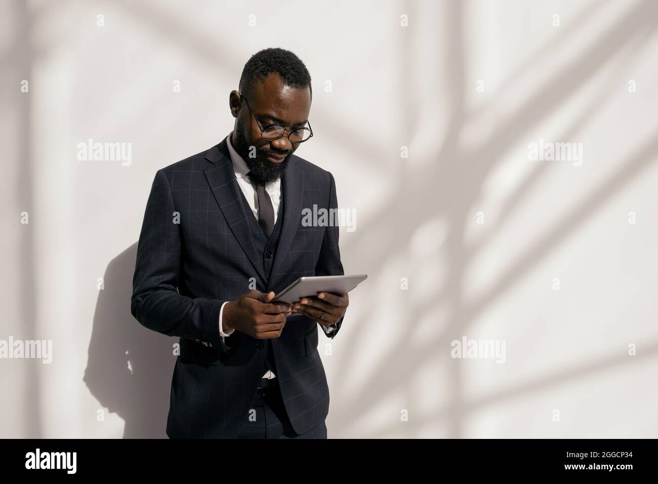 Giovane e serio broker afroamericano con tablet che guarda attraverso le notizie finanziarie contro il muro bianco Foto Stock