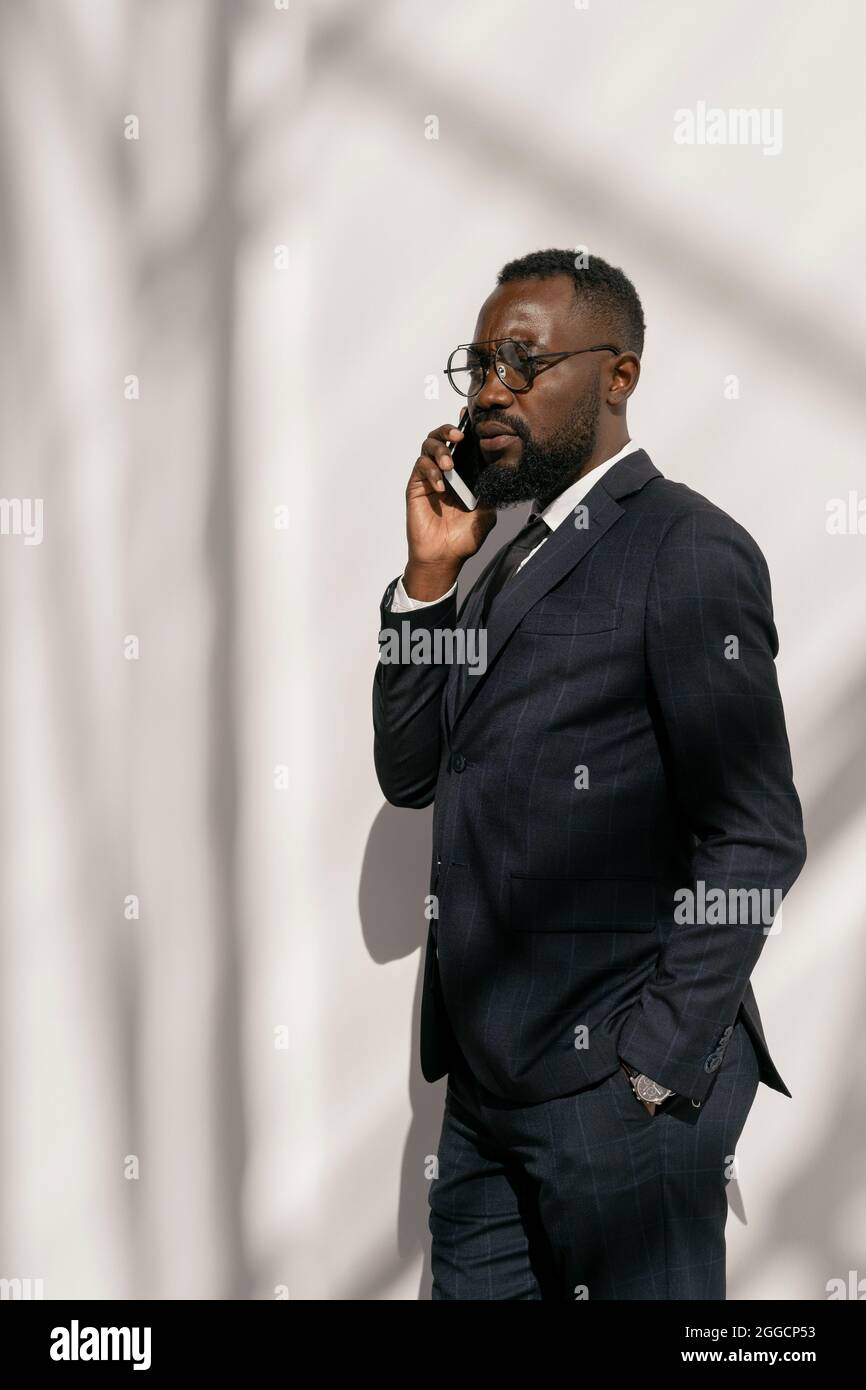 Giovane uomo d'affari contemporaneo in abito elegante parlando al telefono cellulare da parete bianca Foto Stock