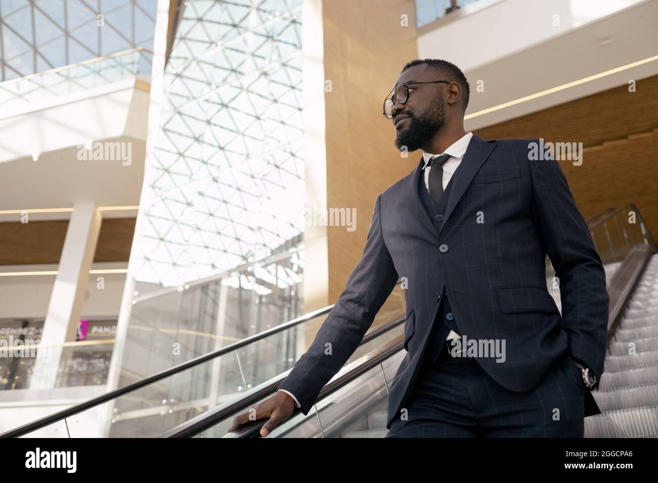 Giovane uomo d'affari fiducioso in tuta in piedi sulla scala mobile mentre si sposta verso il basso nel centro commerciale Foto Stock