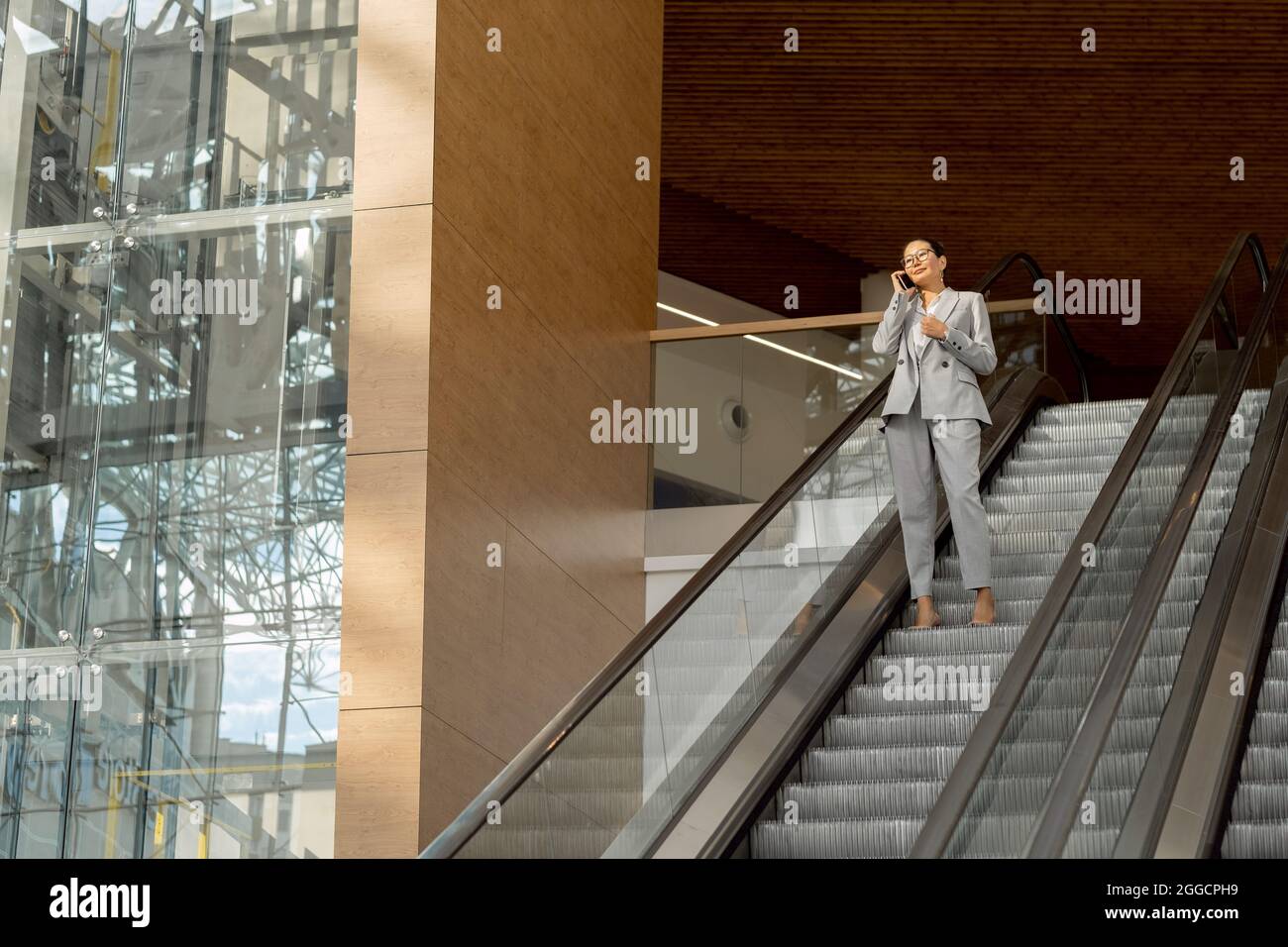 Giovane donna d'affari in abbigliamento formale che parla al telefono cellulare mentre si trova in piedi su scala mobile all'interno di un grande centro commerciale Foto Stock