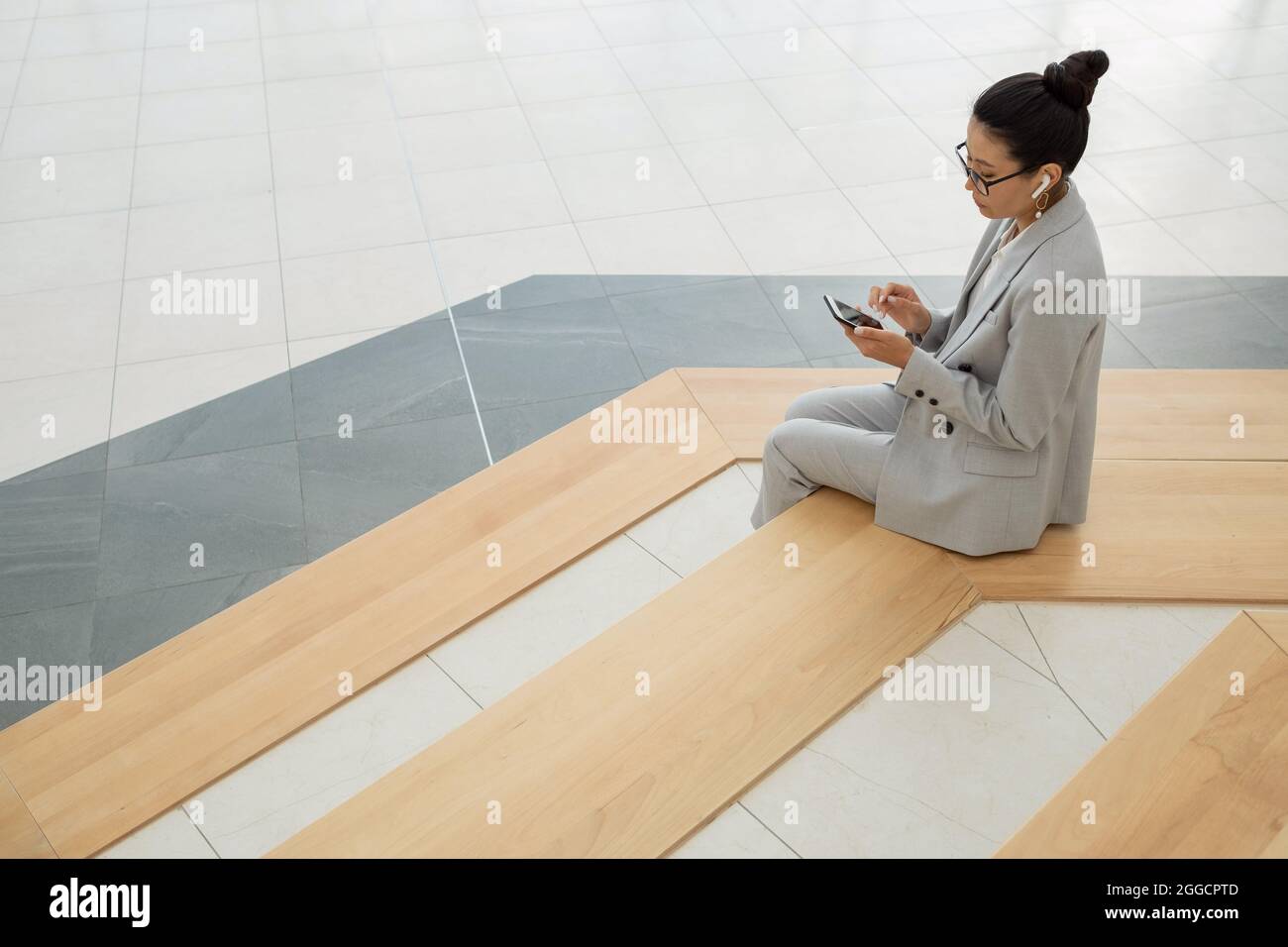 Giovane donna asiatica in abito elegante che ascolta l'audio nello smartphone mentre si siede su grandi scale Foto Stock