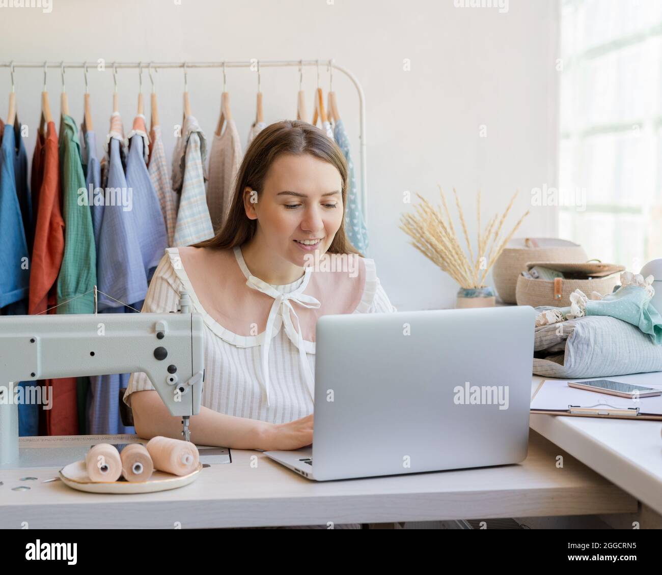 Stilista di moda femminile che crea online abbigliamento design mentre si siede sul posto di lavoro con macchina da cucire Foto Stock