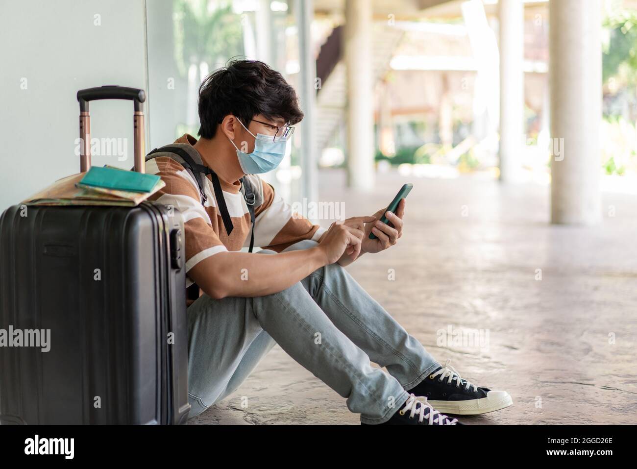 Viaggi. Un uomo che indossa occhiali con una camicia a righe e uno zaino, indossa una maschera, siediti sul pavimento. Utilizzando un telefono durante il viaggio con mappa, notebook sulla su Foto Stock